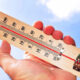 Hand holding a wooden thermometer up to a clear blue sky, showing the temperature with a red liquid line.