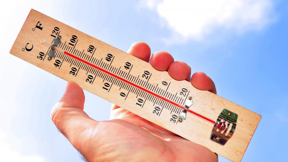 Hand holding a wooden thermometer up to a clear blue sky, showing the temperature with a red liquid line.