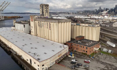 Aerial view of an old industrial complex by the water, with beige silos, a long white-roofed warehouse, and a distant city skyline behind it.
