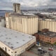 Aerial view of an old industrial complex by the water, with beige silos, a long white-roofed warehouse, and a distant city skyline behind it.