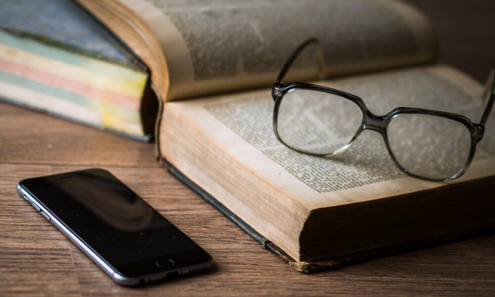 Glasses rest on an open book, with a smartphone on a wooden table nearby.