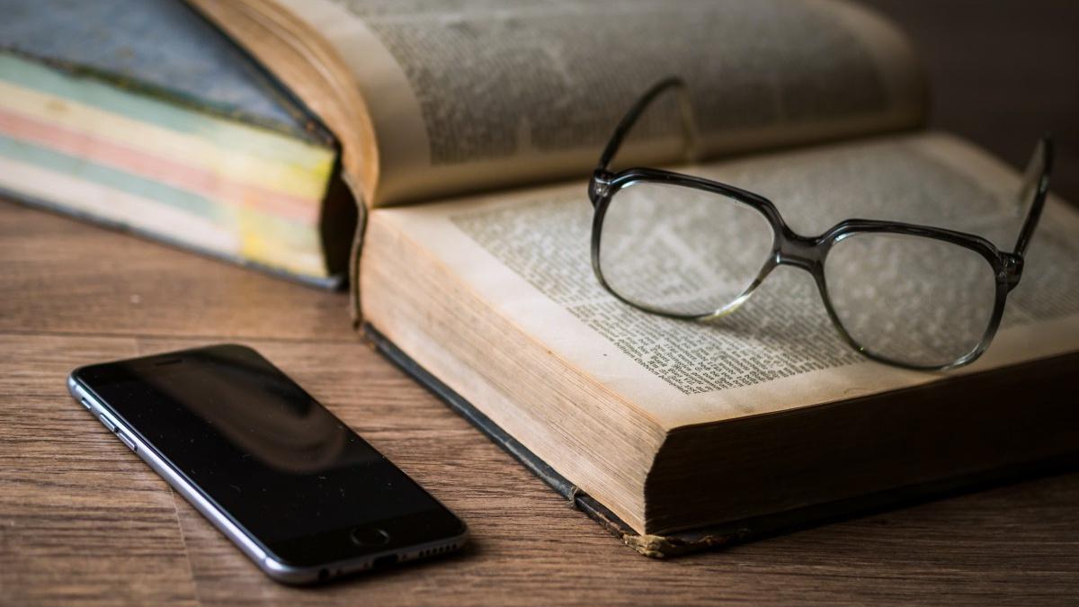 Glasses rest on an open book, with a smartphone on a wooden table nearby.