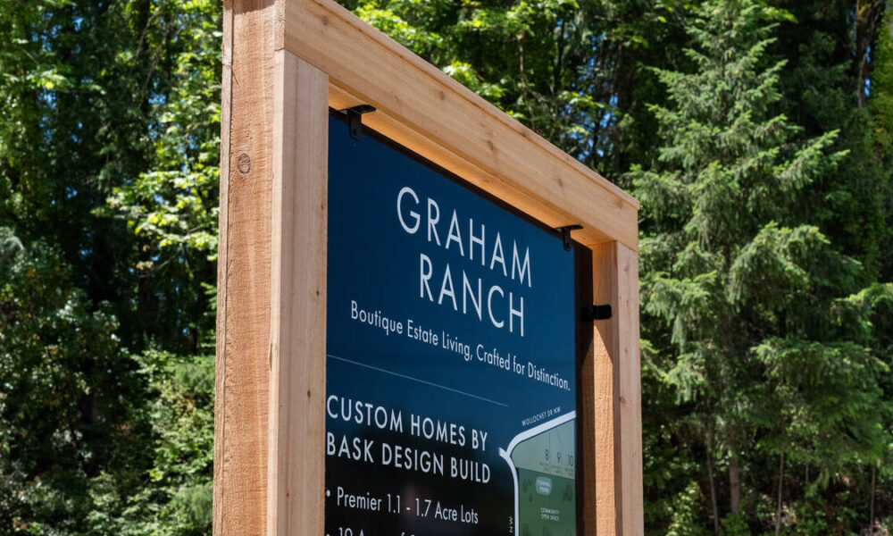 Blue sign for Graham Ranch with text about boutique estate living, framed by a wooden structure among trees in a forest setting, advertising custom homes by Bask Design Build.