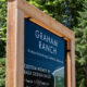 Blue sign for Graham Ranch with text about boutique estate living, framed by a wooden structure among trees in a forest setting, advertising custom homes by Bask Design Build.