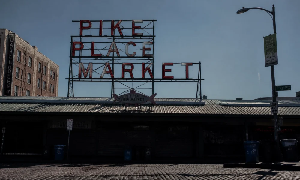 Neon sign reading 'PIKE PLACE MARKET' atop a metal frame over a market building, with a streetlamp and brick buildings nearby on a sunny day.