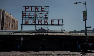 Neon sign reading 'PIKE PLACE MARKET' atop a metal frame over a market building, with a streetlamp and brick buildings nearby on a sunny day.