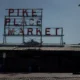 Neon sign reading 'PIKE PLACE MARKET' atop a metal frame over a market building, with a streetlamp and brick buildings nearby on a sunny day.