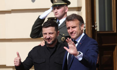Two men pose for a photo outside a building; one gives a thumbs up, the other waves as a uniformed soldier salutes in the background