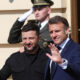 Two men pose for a photo outside a building; one gives a thumbs up, the other waves as a uniformed soldier salutes in the background
