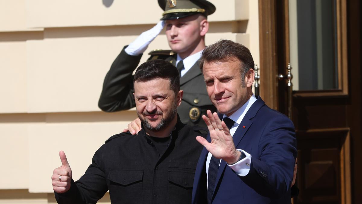 Two men pose for a photo outside a building; one gives a thumbs up, the other waves as a uniformed soldier salutes in the background