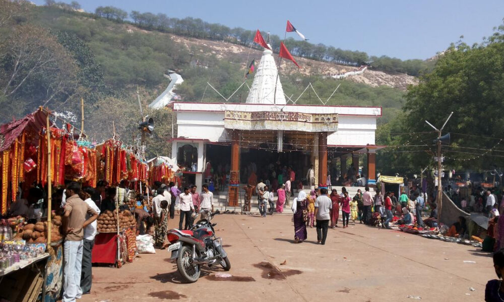 Temple courtyard with a white shrine and red flags, crowded with market stalls and people at an outdoor festival under a clear sky.