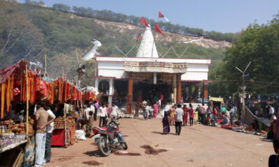 Temple courtyard with a white shrine and red flags, crowded with market stalls and people at an outdoor festival under a clear sky.