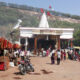 Temple courtyard with a white shrine and red flags, crowded with market stalls and people at an outdoor festival under a clear sky.