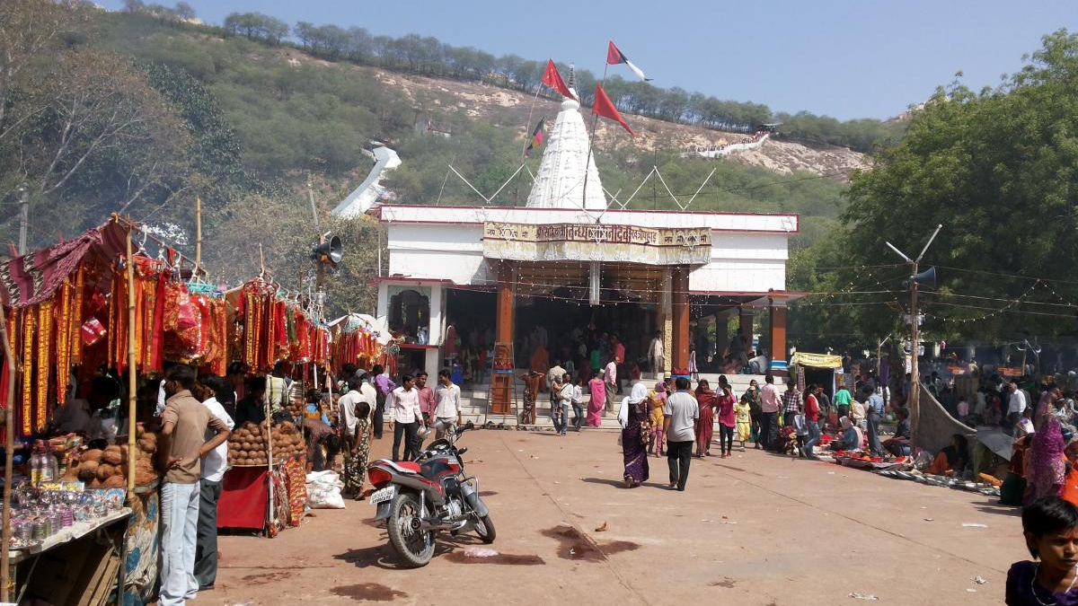Temple courtyard with a white shrine and red flags, crowded with market stalls and people at an outdoor festival under a clear sky.