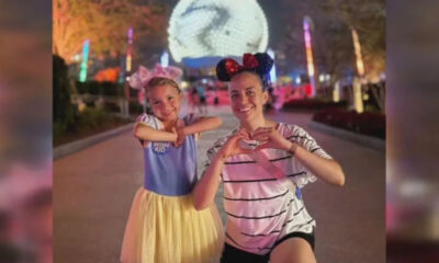 Two smiling people at a nighttime amusement park, making heart shapes with their hands in front of colorful lights and a large glowing decoration behind them.