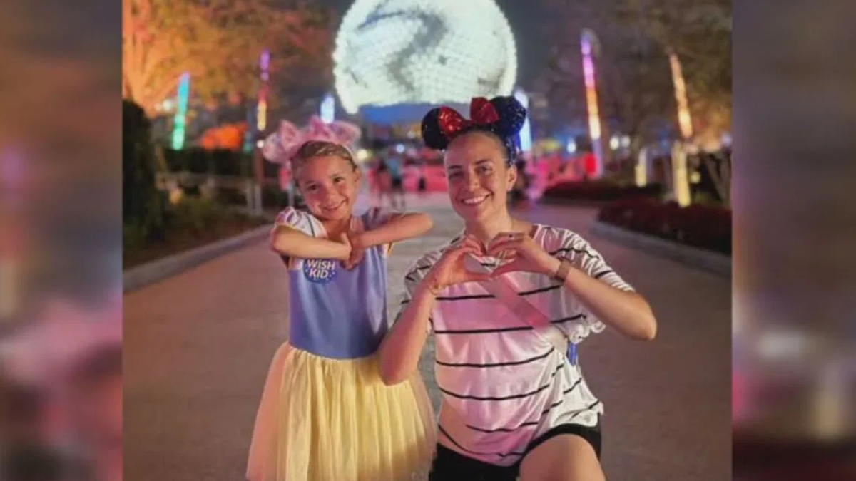 Two smiling people at a nighttime amusement park, making heart shapes with their hands in front of colorful lights and a large glowing decoration behind them.