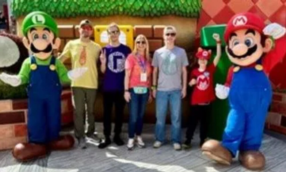 Two life-sized Mario & Luigi mascots flank a group of four visitors posing outdoors in a theme-park-like area.
