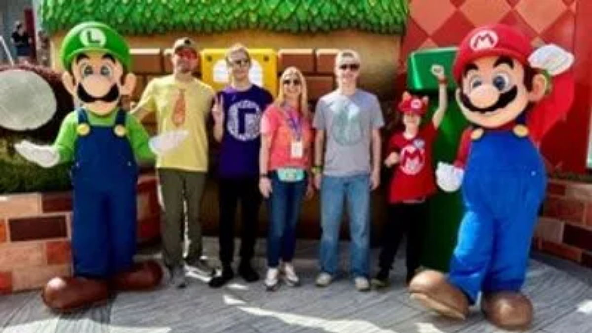 Two life-sized Mario & Luigi mascots flank a group of four visitors posing outdoors in a theme-park-like area.