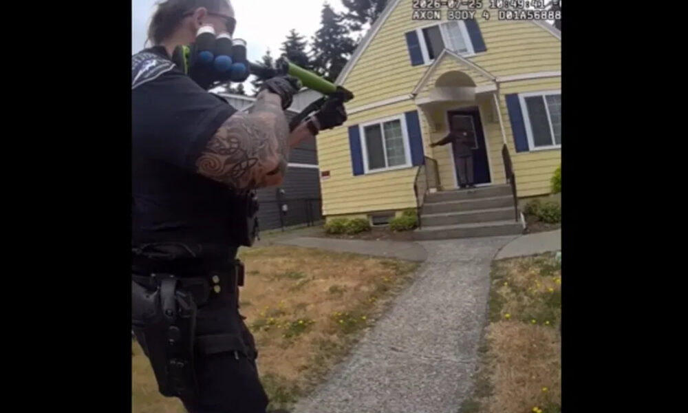 Armed officer aiming a shotgun at the doorway of a yellow house; a person stands on the porch at the top of the steps.