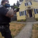 Armed officer aiming a shotgun at the doorway of a yellow house; a person stands on the porch at the top of the steps.