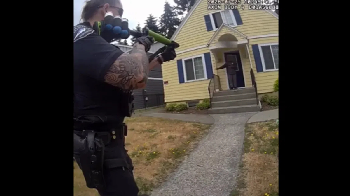 Armed officer aiming a shotgun at the doorway of a yellow house; a person stands on the porch at the top of the steps.