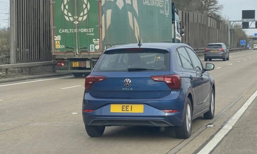 Blue Volkswagen Polo seen from the rear driving on a highway, ahead of a large green cargo truck (UK plate EEI).