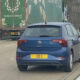 Blue Volkswagen Polo seen from the rear driving on a highway, ahead of a large green cargo truck (UK plate EEI).