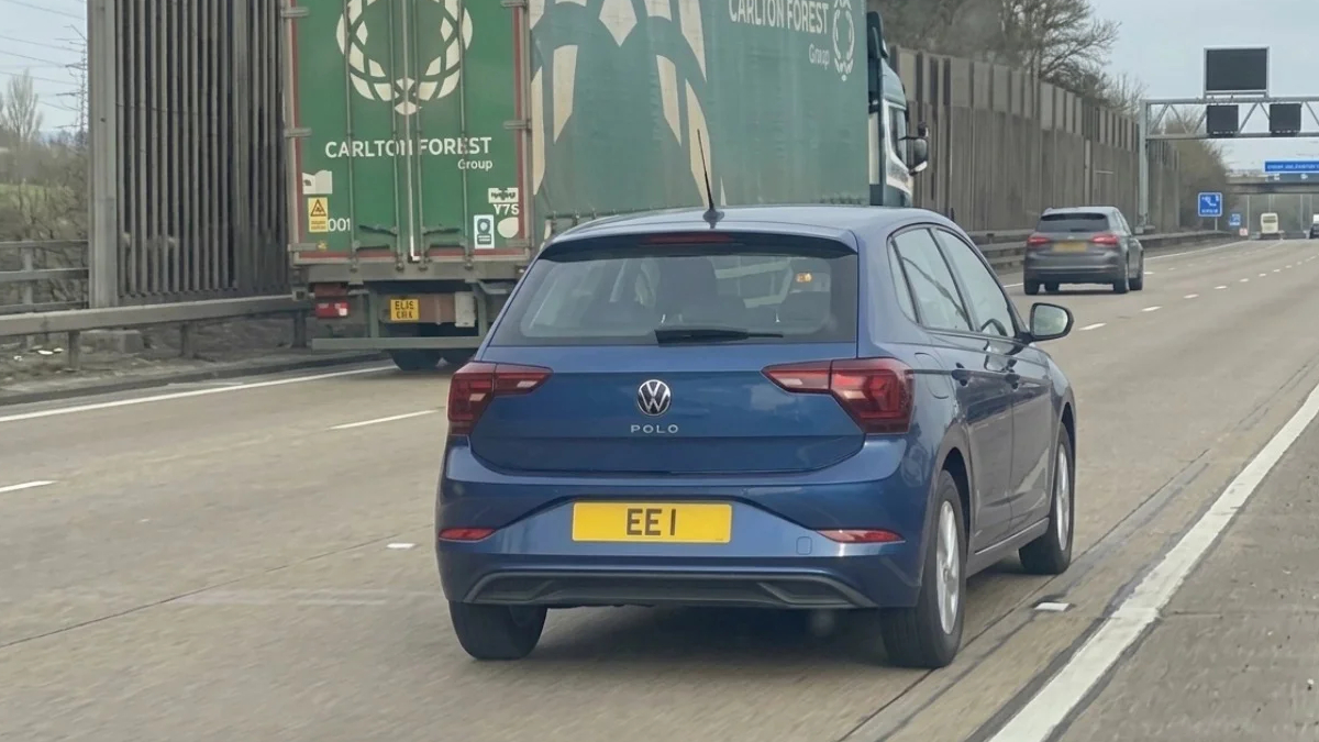 Blue Volkswagen Polo seen from the rear driving on a highway, ahead of a large green cargo truck (UK plate EEI).