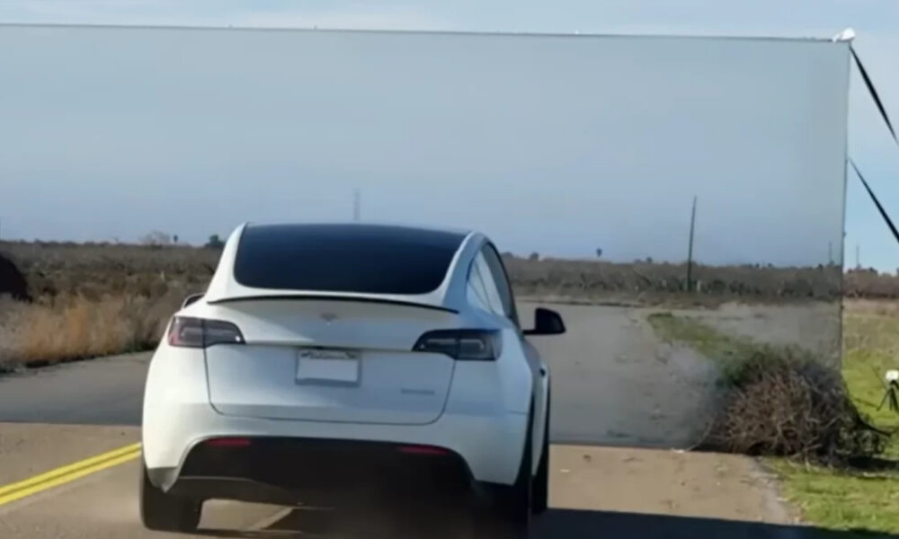 White electric car (Tesla) driving on a rural two-lane road with a large white rectangular backdrop or screen set up on the roadside ahead.