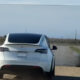 White electric car (Tesla) driving on a rural two-lane road with a large white rectangular backdrop or screen set up on the roadside ahead.