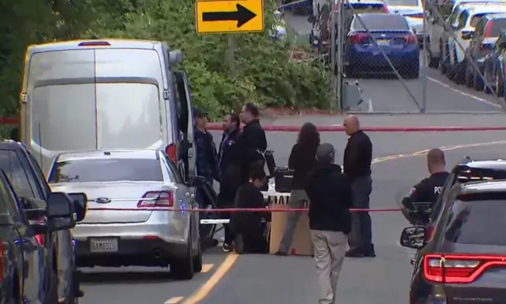 Group of people and police officers stand behind red caution tape around a white van on a city street, indicating an investigation scene.