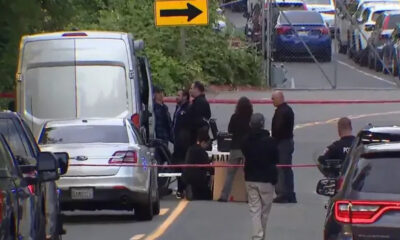 Group of people and police officers stand behind red caution tape around a white van on a city street, indicating an investigation scene.