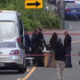 Group of people and police officers stand behind red caution tape around a white van on a city street, indicating an investigation scene.