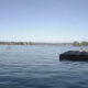 Calm lake scene with a wooden dock on the right and a paddleboarder in the distance, shore lined with trees and houses under a clear blue sky.