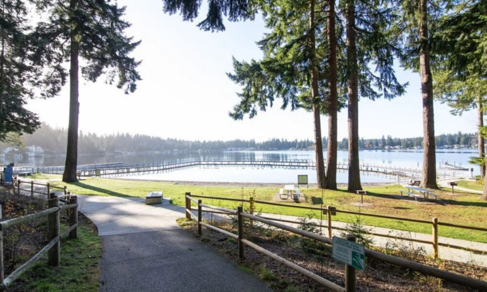 Sunlit lakeside park with a paved path, wooden fence, and tall trees, overlooking a calm lake with a long wooden pier.