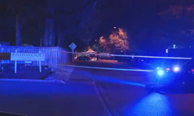 Nighttime police scene with taped-off street, blue emergency lights flashing, and a 'FAIRWOOD' sign on the left.