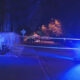 Nighttime police scene with taped-off street, blue emergency lights flashing, and a 'FAIRWOOD' sign on the left.