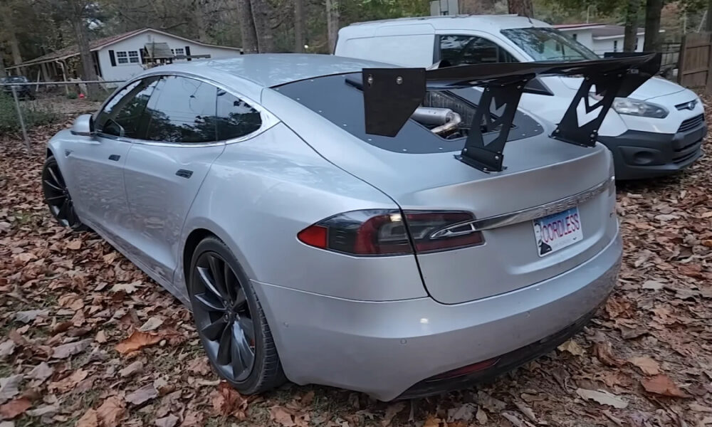 Silver Tesla Model S with a large aftermarket rear wing parked on a leaf-strewn driveway.