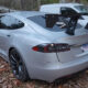 Silver Tesla Model S with a large aftermarket rear wing parked on a leaf-strewn driveway.