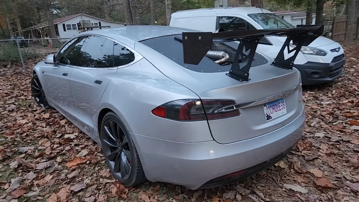 Silver Tesla Model S with a large aftermarket rear wing parked on a leaf-strewn driveway.