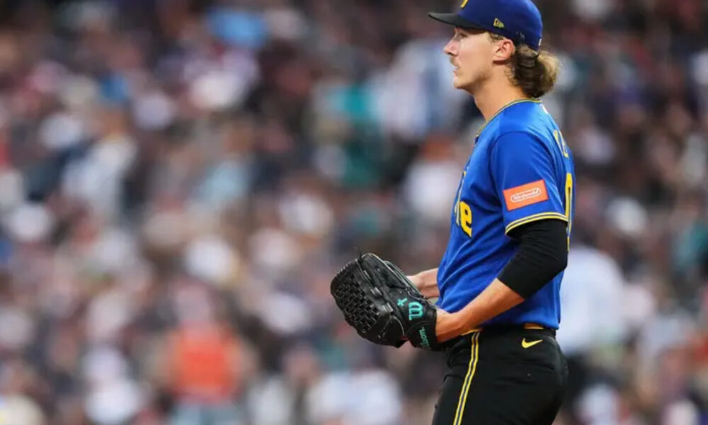 Profile view of a baseball player in a blue jersey and cap, holding a glove as he stands on the field with a blurred crowd in the background.