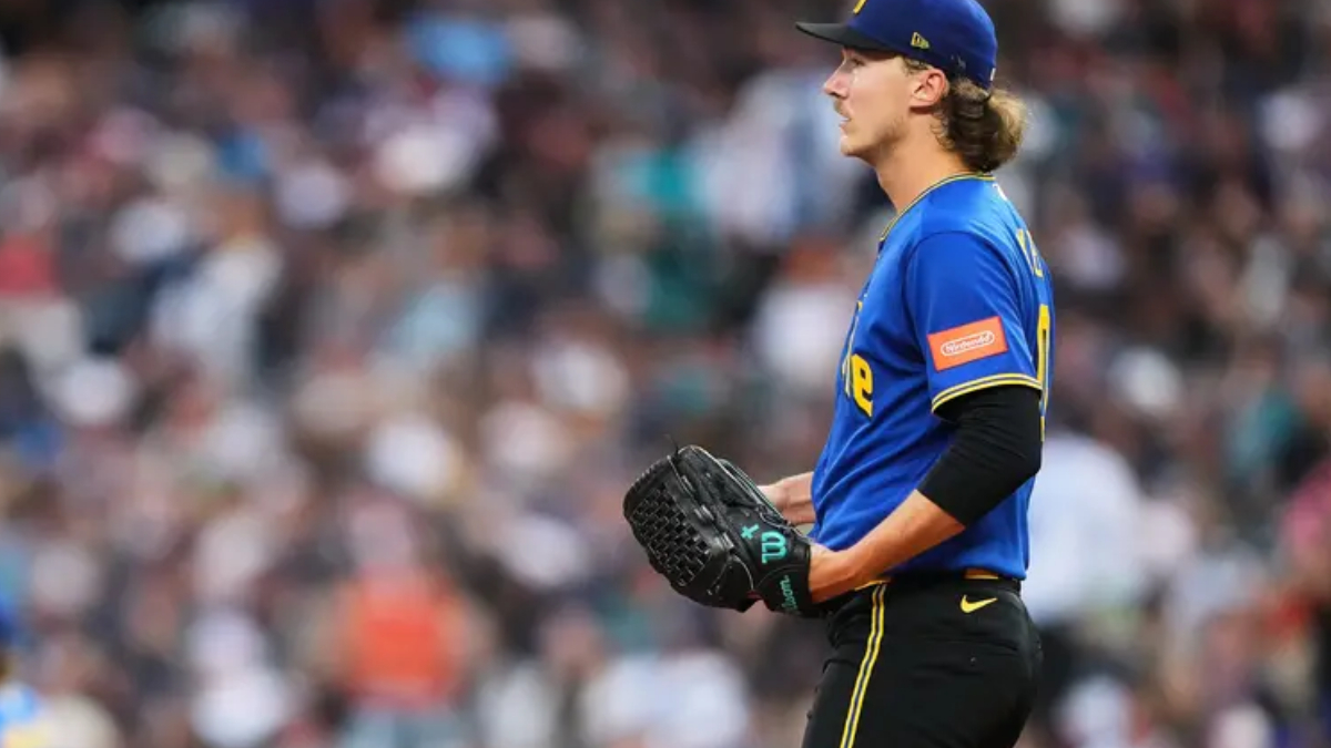 Profile view of a baseball player in a blue jersey and cap, holding a glove as he stands on the field with a blurred crowd in the background.