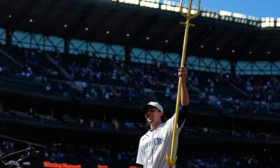 Mariners player in white uniform raises a tall golden ceremonial spear on the field with a packed stadium in the background.