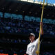Mariners player in white uniform raises a tall golden ceremonial spear on the field with a packed stadium in the background.