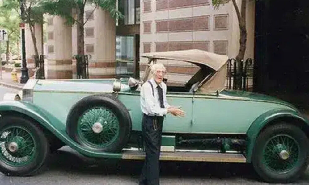 Older man with white hair in a white shirt and tie stands beside a mint-green vintage convertible on a city street.