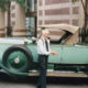 Older man with white hair in a white shirt and tie stands beside a mint-green vintage convertible on a city street.