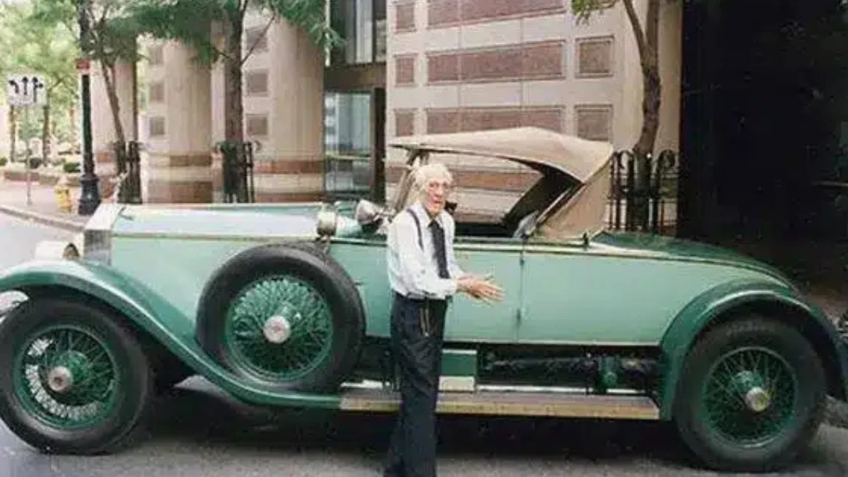 Older man with white hair in a white shirt and tie stands beside a mint-green vintage convertible on a city street.