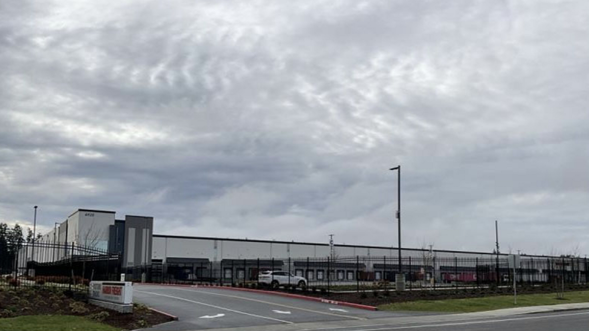 Industrial warehouse complex behind a tall black fence, with a curved road and overcast sky.
