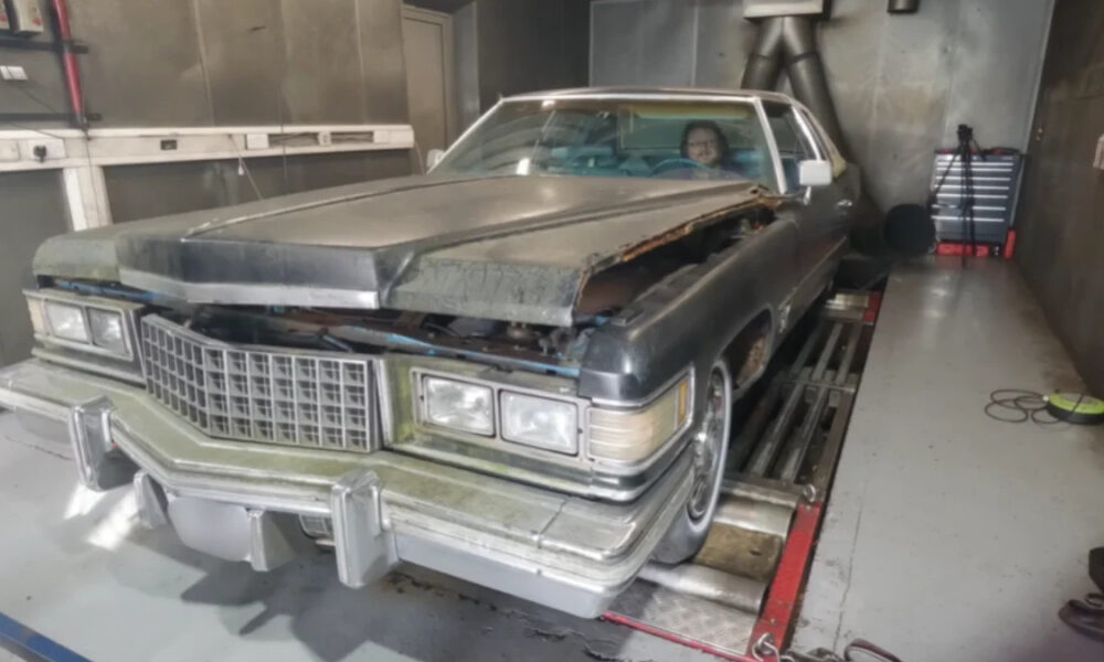 Dusty vintage Cadillac with hood open and front-end damage in a workshop; a person sits in the driver's seat.