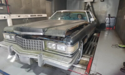 Dusty vintage Cadillac with hood open and front-end damage in a workshop; a person sits in the driver's seat.